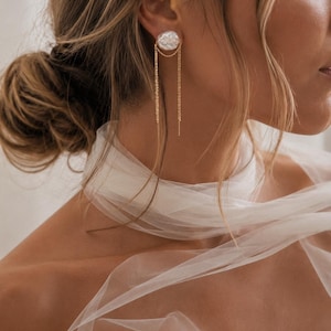 May include: A gold stud earring featuring a textured, irregular white circular disc. Two delicate gold chains hang from the base of the disc, draping downwards to create a layered effect. The earring is worn on the earlobe, showcasing a metallic finish.
