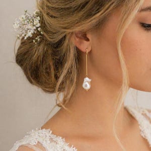 May include: A close-up of a woman wearing a gold earring with a white pearl. The earring hangs from a thin gold bar. The woman has blonde hair styled in a bun with small white flowers. She is wearing a white lace dress.