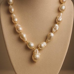 Peut inclure: Un collier de perles de couleur crème exposé sur un mannequin. Le collier est composé de perles de forme irrégulière enfilées avec de petites perles dorées. Le pendentif est une perle unique, plus grande.