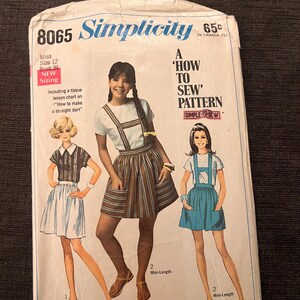 Op de afbeelding: Vintage Simplicity naaipatroon, maat 12, met illustraties van drie verschillende mini-outfits. Het patroon bevat een leskaart over het maken van een rechte dart. De omslag is wit met blauwe en rode tekst.