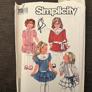 May include: A Simplicity sewing pattern with illustrations of children's dresses. The pattern shows various dress designs, including those with ruffles, bows, and Peter Pan collars. The pattern is in a white envelope, with handwritten notes.