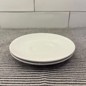 May include: Two white ceramic saucers stacked. Each saucer features a ridged, radial design around the edge. The saucers rest on a striped, grey and white fabric, with a white tiled background.