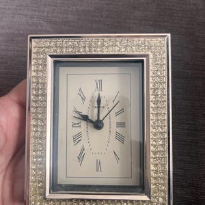 Puede incluir: Un reloj de cuarzo rectangular con un marco plateado incrustado con pequeños cristales transparentes. La esfera del reloj es blanquecina con números romanos y manecillas negras. El reloj muestra la hora aproximadamente a las 11:45.