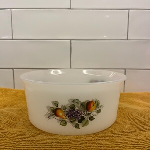 May include: A white, round bowl with a fruit design featuring pears, grapes, and an apple. The bowl is sitting on a yellow towel. The fruit design is in shades of green, purple, yellow, and red.