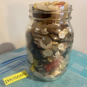May include: A glass jar packed with an assortment of buttons in various colours and materials. Buttons include white, brown, black, and iridescent types. A yellow sticky note with the word "IRRISDESENTS" is visible.