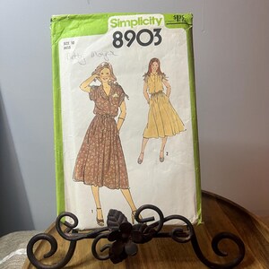 Op de afbeelding: Een vintage Simplicity 8903 naaipatroon met illustraties van twee jurken. De patroonhoes is groen en wit, met maat 10 Miss genoteerd. Het ontwerp omvat een bruine jurk met bloemenprint en een gele jurk.