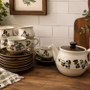May include: A ceramic tea set featuring a teapot and multiple cups with saucers. The pieces are cream-coloured with a hand-painted floral motif of small green, brown, and blue leaves. The teapot lid and saucers are a solid, glossy dark brown glaze.