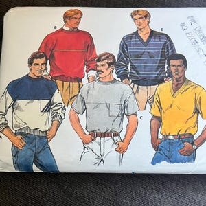 May include: A sewing pattern envelope featuring illustrations of five different tops. The designs include a red crewneck sweater, a blue striped V-neck sweater, a color-blocked crewneck sweatshirt, a light gray short-sleeve t-shirt, and a yellow V-neck t-shirt.