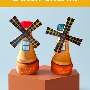 Puede incluir: Dos saleros y pimenteros de madera con forma de molinos de viento. Los saleros son amarillos con detalles rojos y azules, y aspas negras y amarillas. El salero tiene escrito "Salt", y el pimentero "Pepper". El texto en la parte superior dice "Add a Pinch of Dutch Charm."