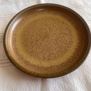 May include: A round ceramic plate with a speckled, golden-brown centre and a darker brown rim. The plate has a slightly raised edge and a matte finish. The plate is likely for serving food.