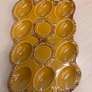 May include: A vintage, mustard-yellow ceramic egg holder with a glossy finish. The tray has 12 oval-shaped egg compartments and two round compartments, with ornate, raised detailing around the edges and between the egg slots. A small hole is at the base.