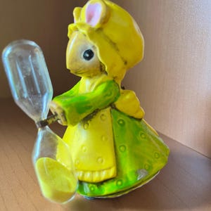 May include: A small figurine of a mouse wearing a yellow bonnet and a green dress with a yellow apron. The mouse is holding a clear hourglass filled with yellow sand. The figurine features painted details including pink inner ears and black eyes.