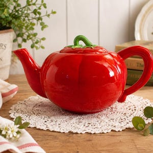Puede incluir: Una tetera de cerámica roja brillante con forma de tomate, que presenta un cuerpo redondeado con sutiles estrías verticales, un pico curvo, un asa resistente y una tapa extraíble rematada con un pequeño tallo verde esculpido.