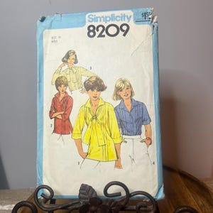 Peut inclure: Patron de couture Simplicity 8209 vintage avec des illustrations de chemisiers rouges, jaunes et bleus. Le patron est pour une taille 10 Miss. La couverture est bleu clair avec le logo Simplicity et le prix.
