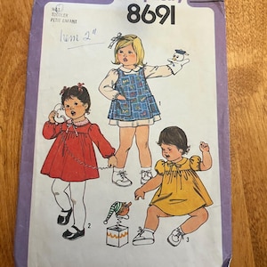 Może przedstawiać: Vintage wzór szycia Simplicity 8691 dla sukienek dla małych dzieci. Okładka wzoru zawiera ilustracje trzech małych dzieci w różnych stylach sukienek. Wzór zawiera tekst "TODDLER PETIT ENFANT" i "hem 2".