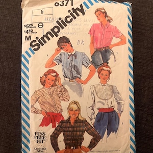 May include: A vintage Simplicity sewing pattern envelope with illustrations of women's blouses. The envelope is white with blue stripes and the Simplicity logo. The text "Fuss-Free Fit" is visible.
