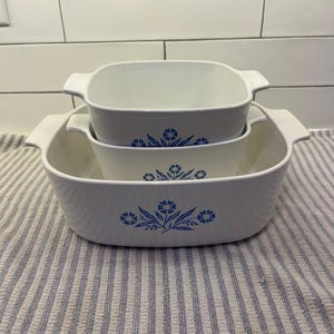 Może przedstawiać: Stos trzech białych ceramicznych naczyń do pieczenia z niebieskimi wzorami kwiatowymi. Naczynia są prostokątne z zaokrąglonymi rogami i uchwytami. Wzór kwiatowy to stylizowany niebieski kwiat z liśćmi.