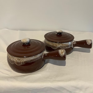 Puede incluir: Dos cuencos de sopa de cerámica marrón con tapas y asas. Los cuencos tienen un acabado brillante con un borde y un pomo de tapa más claros y moteados. Las asas están integradas en el diseño del cuenco, ofreciendo una estética clásica y funcional.