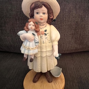 Könnte beinhalten: Porzellanfigur eines jungen Mädchens in einem cremefarbenen Kleid und einem breitkrempigen Hut, das eine Puppe hält. Die Figur steht auf einem Holzsockel und weist zarte, bemalte Details auf, darunter florale Akzente und eine kleine Schüssel.