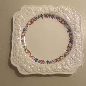 Op de afbeelding: Witte vierkante plaat met een opstaande, sierlijke rand. Het midden heeft een bloemen- en fruitontwerp in een cirkelvormig patroon. De plaat heeft een geschulpte rand en een glanzende afwerking.