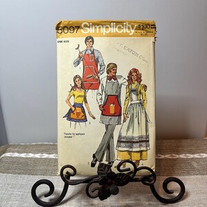Puede incluir: Un patrón de costura Simplicity vintage, número 5097, con ilustraciones de delantales. El patrón presenta delantales en rojo y blanco. El patrón se muestra en un soporte decorativo de metal negro.