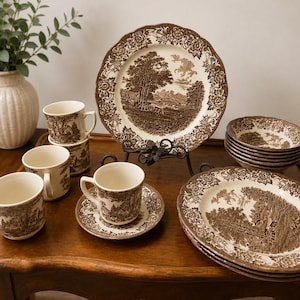 May include: A collection of cream-colored ceramic dinnerware featuring a brown transferware pattern of rural landscapes and floral borders. The set includes several plates, a stack of bowls, multiple mugs, and a saucer, all displaying a consistent pastoral scene.