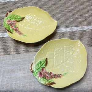 以下が含まれることがあります： 淡い黄色の釉薬を施した、葉の形をした2つのセラミック皿。各皿には葉脈の隆起模様があり、緑の葉、ピンクの花、茶色のアクセントで装飾されています。お皿には小さな取っ手が付いています。