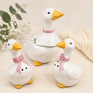 May include: A ceramic condiment set featuring a central goose-shaped lidded jar with a silver spoon, accompanied by two smaller goose-shaped shakers. Each white goose has a yellow beak, yellow feet, and a pink ribbon tied around its neck.