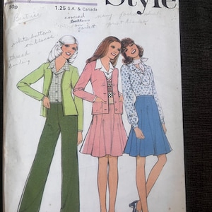 May include: A vintage sewing pattern cover with illustrations of three women in 1970s attire. The cover displays the word "Style" and handwritten notes. The outfits include a green trouser suit, a pink skirt suit, and a blue skirt with a patterned blouse.