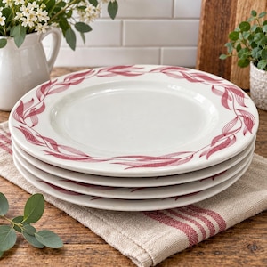 May include: A stack of four white ceramic dinner plates featuring a continuous border pattern of stylized, thin, dark red leaves. The plates have a smooth, glossy finish and a wide, flat rim, with the red leaf design encircling the entire edge.
