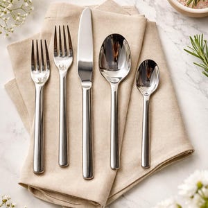 May include: A set of polished stainless steel cutlery, comprising forks, a knife, and spoons, presented on a beige linen napkin. The silverware reflects light, with white flowers and a small bowl of pink salt adding to the scene.