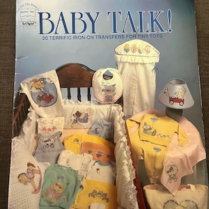 May include: A selection of baby clothes, bibs, and a lampshade, all decorated with cartoon bear motifs. The items are displayed in a crib-like arrangement, with a book titled "BABY TALK!" in the background. The color scheme features soft pastels and bright primary colours.