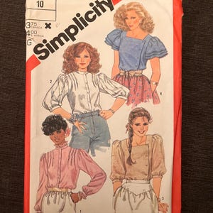 May include: A vintage Simplicity sewing pattern showing illustrations of women's blouses. The pattern includes various blouse styles with details like puffed sleeves, button closures, and high collars. The cover is red and white with the Simplicity logo.