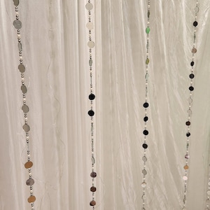 Puede incluir: Varias hebras verticales de cuentas plateadas, negras e iridiscentes en forma de disco cuelgan frente a una cortina blanca y transparente. Las cuentas están conectadas por pequeñas cuentas plateadas, creando un efecto decorativo y brillante.