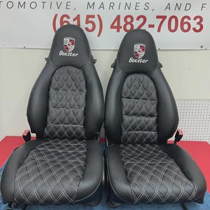 May include: A pair of black leather car seats featuring diamond stitching in white. Each seat has a Porsche logo and the word "Boxster" embroidered on the headrest. The seats are displayed on a red surface, with a white and red sign in the background.
