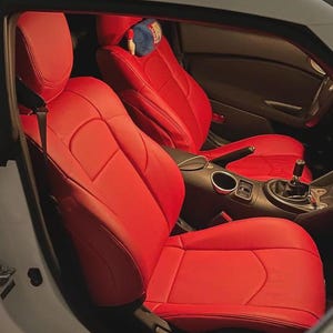 May include: Bright red leather car seats. The seats are in a car with a white exterior. The interior features a black dashboard and center console. A small blue pillow with a design is on the headrest.