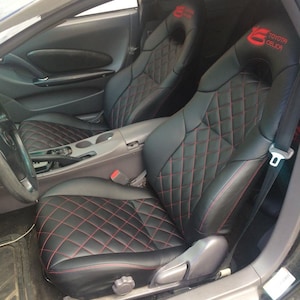 Puede incluir: Asientos de coche de cuero negro con costuras de diamante rojas. Los asientos tienen las palabras "Toyota Celica" bordadas en los reposacabezas.