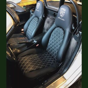 Op de afbeelding: Zwarte leren autostoelen met witte diamantstiksels. De hoofdsteunen tonen het Porsche-embleem en het woord "Boxster". De stoelen bevinden zich in een zilverkleurig auto-interieur.