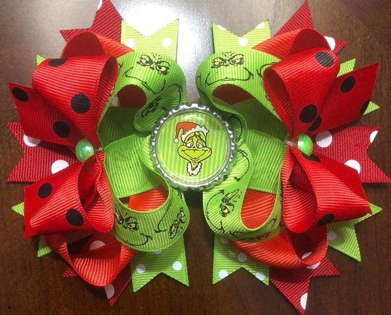 May include: A festive hair bow featuring red and green ribbons with black polka dots and a Grinch-themed bottle cap center. The bow is adorned with a green bead and is perfect for holiday celebrations.