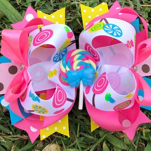 May include: A pink and white candy-themed hair bow with a blue and yellow lollipop accent. The bow is made of multiple layers of ribbon with different candy patterns.
