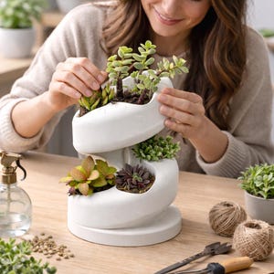 May include: A white, spiral-shaped planter filled with various succulents. The planter has three levels, each holding different types of green and reddish-brown succulents. A person is tending to the plants. Gardening tools and twine are on the wooden surface.