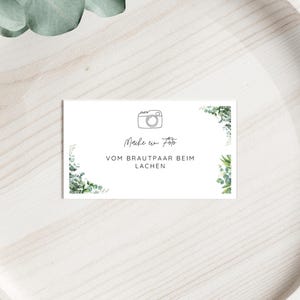 May include: A white card with the text "Make a photo of the bride and groom laughing" and a camera illustration. The card is decorated with green eucalyptus leaves and is placed on a light wooden surface.