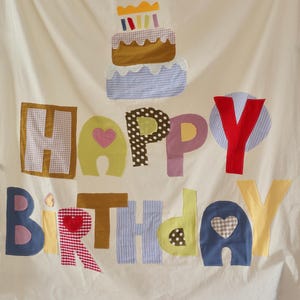 May include: A cream-colored fabric banner featuring a multi-colored fabric applique of a birthday cake at the top. Below the cake, the words HAPPY BIRTHDAY are spelled out in large, individual fabric letters with various patterns and colors.