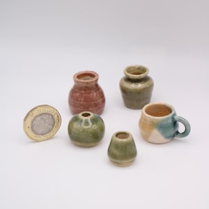 May include: A selection of small ceramic pots and a miniature mug, in shades of brown, green, and blue. A one-pound coin is included to show the scale of the pottery. The pieces are on a white background.