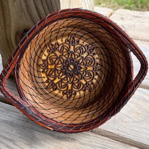 May include: Handwoven basket with a mandala design. The basket features a spiral pattern with shades of brown, orange, and tan. The center of the basket has a detailed mandala design. The basket has two handles.