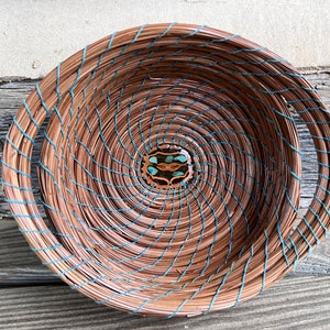 May include: A woven basket with a circular shape and two handles. The basket is made of brown and blue materials and has a decorative center piece with turquoise accents.