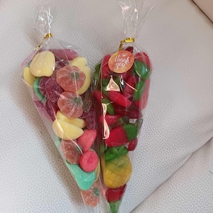 May include: Two cellophane bags filled with colourful gummy sweets shaped like fruits. One bag contains red, green, and yellow sweets, and the other has red, pink, yellow, and green sweets. One bag has a small orange sticker that says "thank you".
