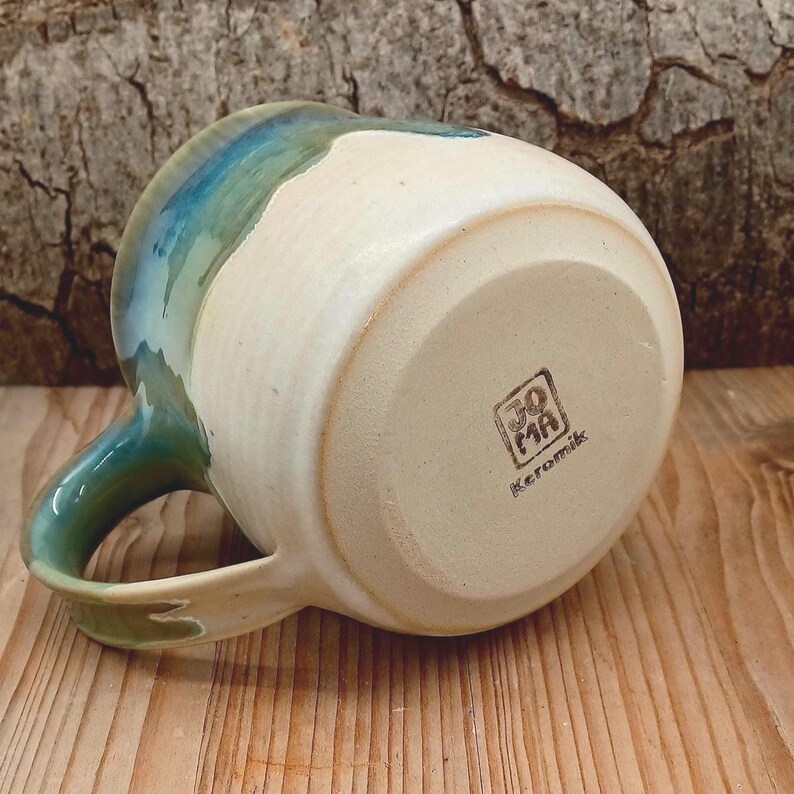 Puede incluir: Una taza de cer&aacute;mica hecha a mano con un cuerpo color crema y un borde y asa esmaltados en azul verdoso y azul. La parte inferior de la taza tiene un logotipo estampado que dice "JO MA Keramik". La taza est&aacute; sobre un fondo de madera.