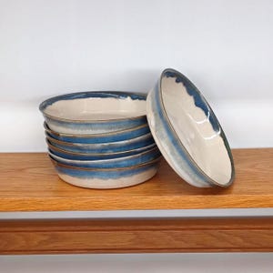 May include: A stack of six ceramic bowls with a cream-colored interior and a blue and white glazed exterior. The bowls are displayed on a wooden shelf against a white background. The bowls have a simple, elegant design.