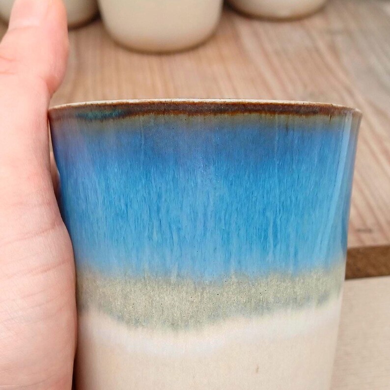 Op de afbeelding: Een keramische beker met een gradi&euml;ntontwerp. De bovenkant van de beker is diep bruin, overgaand in een levendig blauw, dan een lichtgroen en tenslotte een cr&egrave;mewit aan de basis. De beker wordt door een hand vastgehouden.